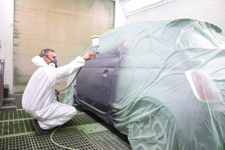 Mitarbeiter in Schutzanzug lackiert eine abgeklemmte Fahrzeugseite mit der Spritzpistole in der Lackierkabine.