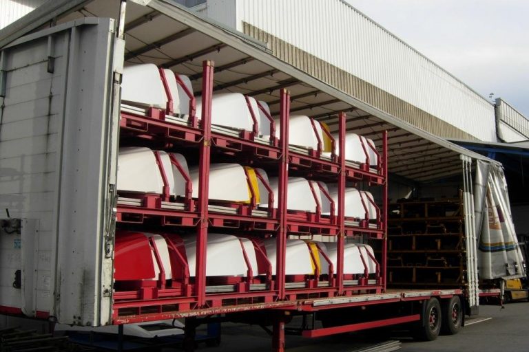 Offener LKW beladen mit gestapelten, lackierten Fahrzeuganbauteilen in roten Transportgestellen.