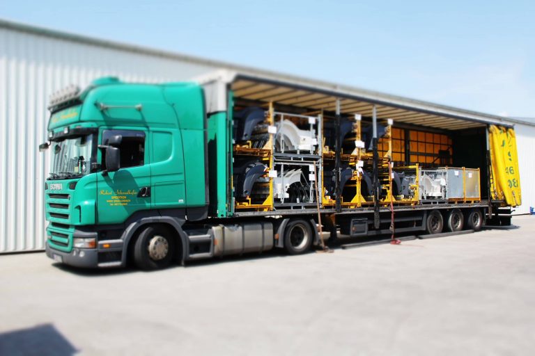 Grüner LKW mit geöffnetem Auflieger, beladen mit lackierten Fahrzeugteilen in gelben Transportgestellen.